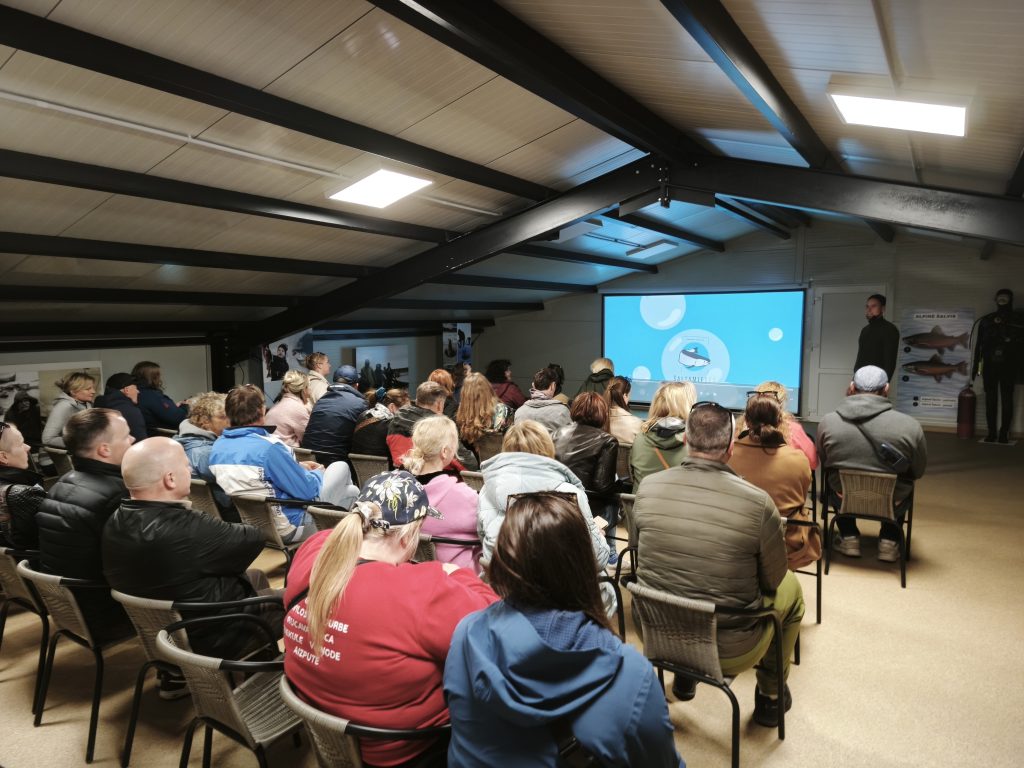 Audience attending a presentation in a conference room with a screen displaying information, featuring a speaker and various educational materials on the walls.