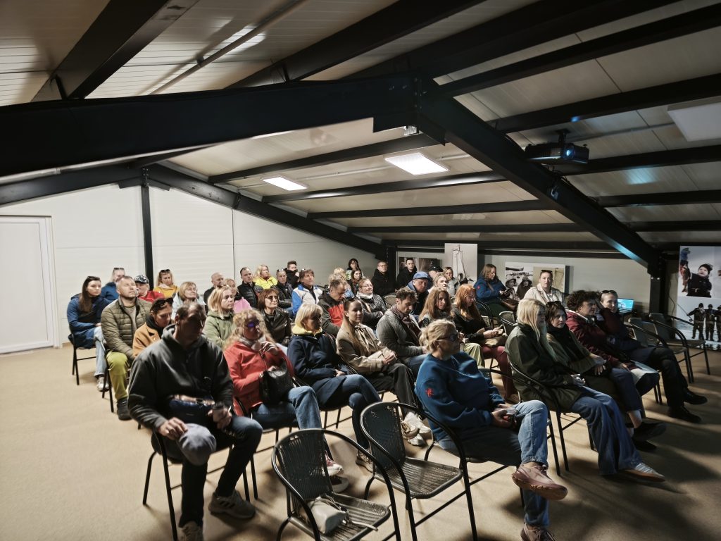 Audience gathered in a modern indoor space for a presentation or event, seated in rows of chairs, with a projector and display visible in the background. The setting features a high ceiling with exposed beams, creating an engaging atmosphere for learning or discussion.