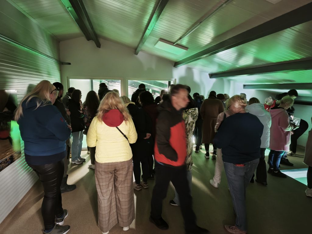 Crowd of people gathered in a dimly lit room with green lighting, engaged in conversation and observing the surroundings. The scene captures a diverse group with various clothing styles, creating a lively atmosphere.