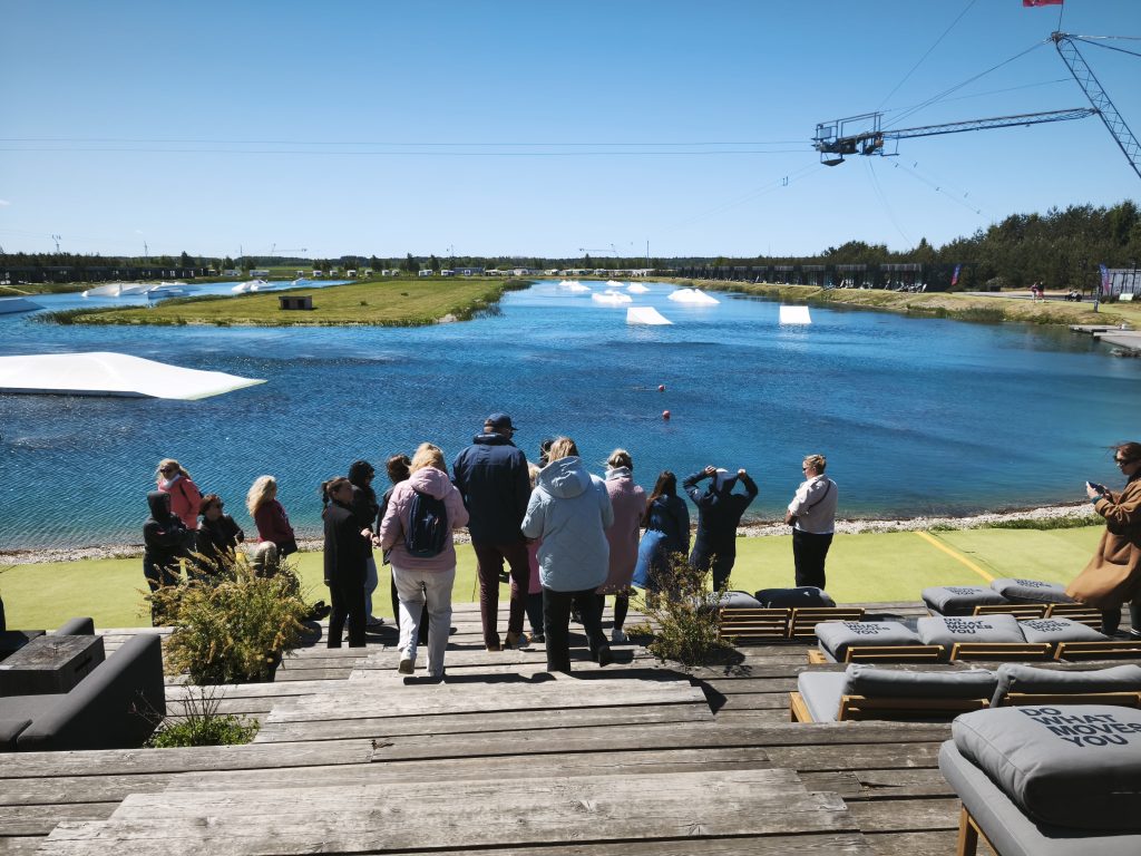 Group of people gathered by a waterfront at a wakeboarding facility, with a scenic view of blue water and wakeboarding ramps in the background. Clear skies and greenery enhance the outdoor experience.