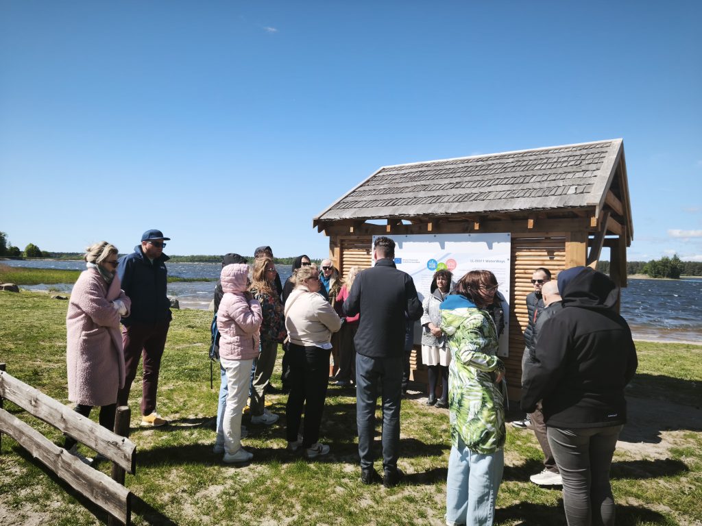 Group of people gathered near a wooden information kiosk by a lake on a sunny day, discussing local waterway conservation and education. Green grass and water in the background enhance the scenic view.