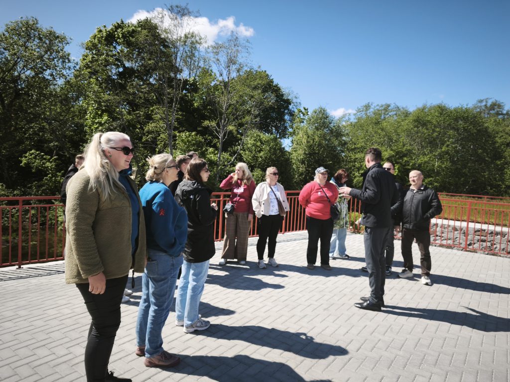 Group of tourists engaged in a guided tour outdoors, standing on a paved area surrounded by greenery and a clear blue sky. The tour guide, dressed in black, is speaking to the attentive audience, which includes men and women of various ages.
