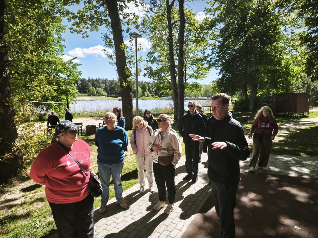 Group of people listening to a speaker during an outdoor tour by a lake, surrounded by trees and greenery on a sunny day.