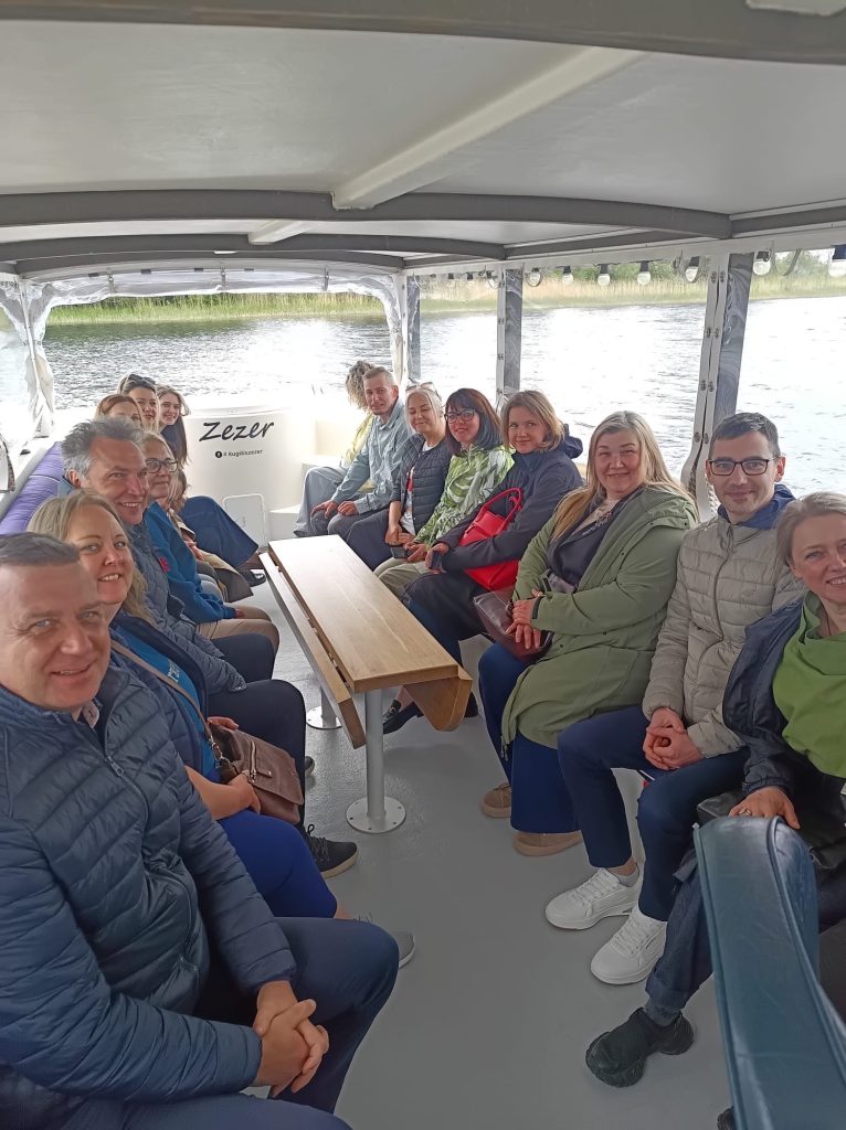 Group of people enjoying a boat trip on a river, seated comfortably inside a covered boat with a wooden table. The scene captures a diverse group, smiling and engaging with one another, surrounded by scenic views of water and greenery. Group of people enjoying a boat trip on a river, seated comfortably inside a covered boat with a wooden table. The scene captures a diverse group, smiling and engaging with one another, surrounded by scenic views of water and greenery.
