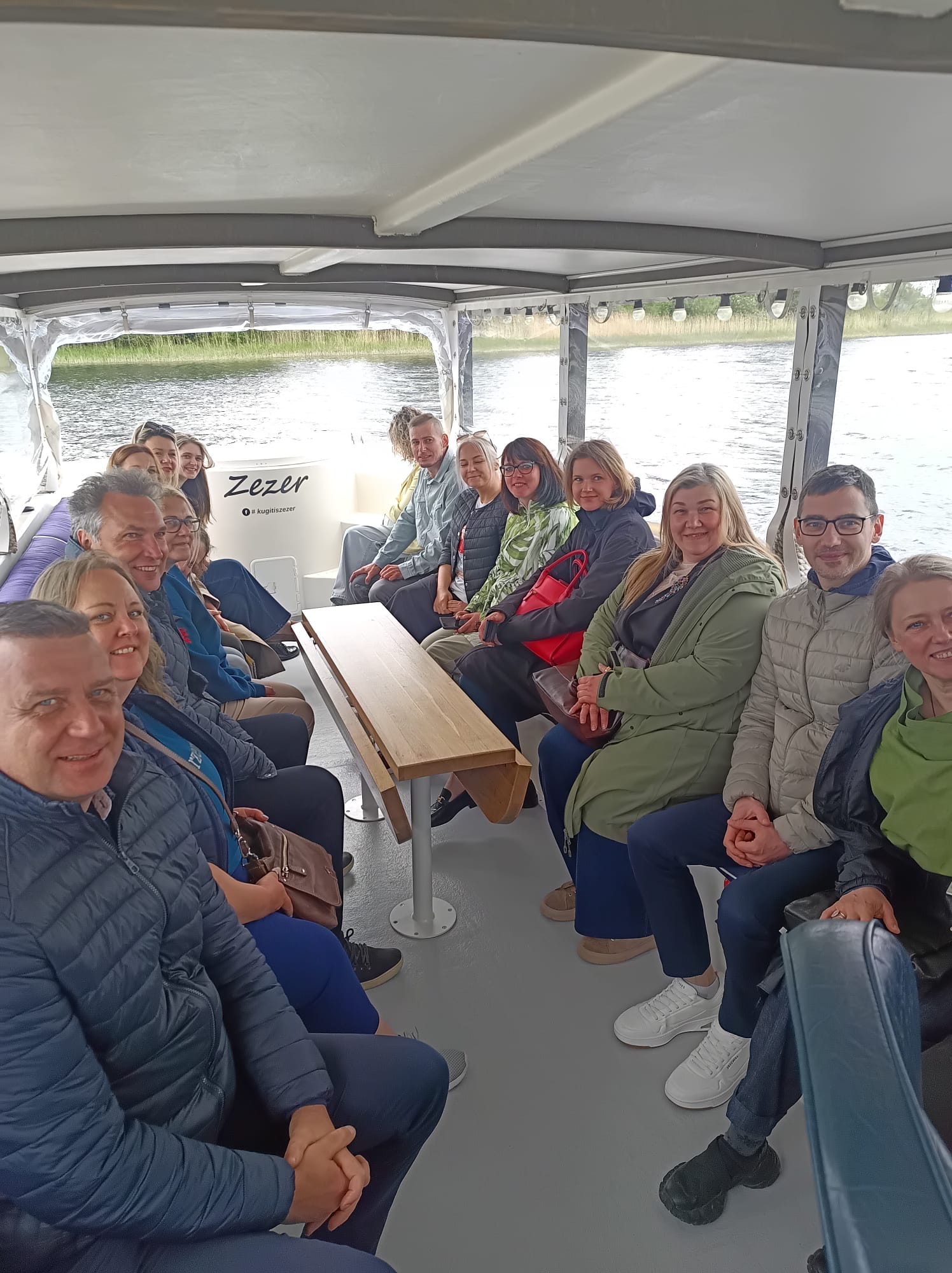 Group of people enjoying a boat tour on the water, seated on a small boat with a wooden table in the center, surrounded by lush greenery. The boat is named "Zezer." Group of people enjoying a boat tour on the water, seated on a small boat with a wooden table in the center, surrounded by lush greenery. The boat is named "Zezer."