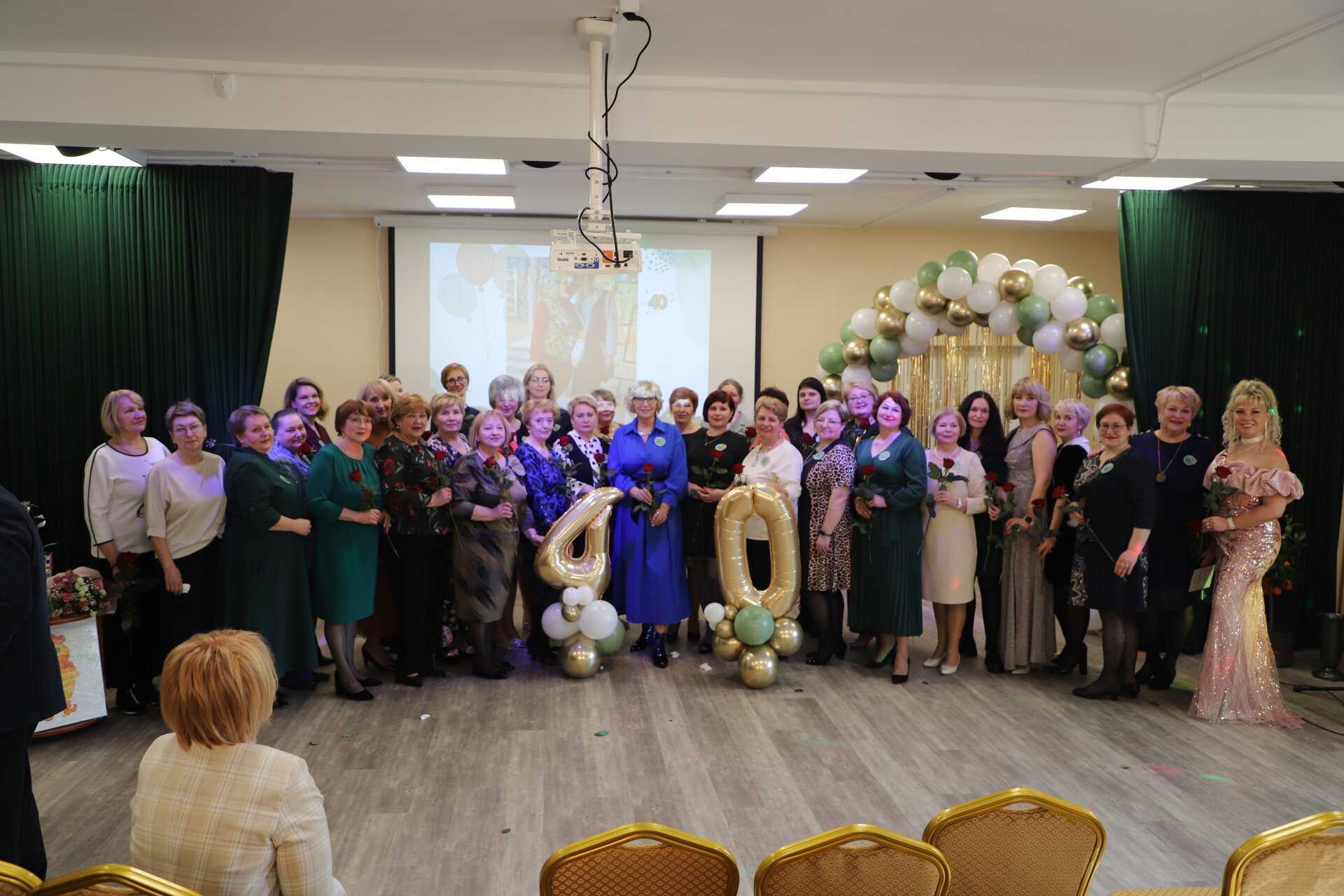 Group of women celebrating a 40th anniversary event, standing together with roses in hand, in a decorated venue featuring balloons and a projection screen in the background.