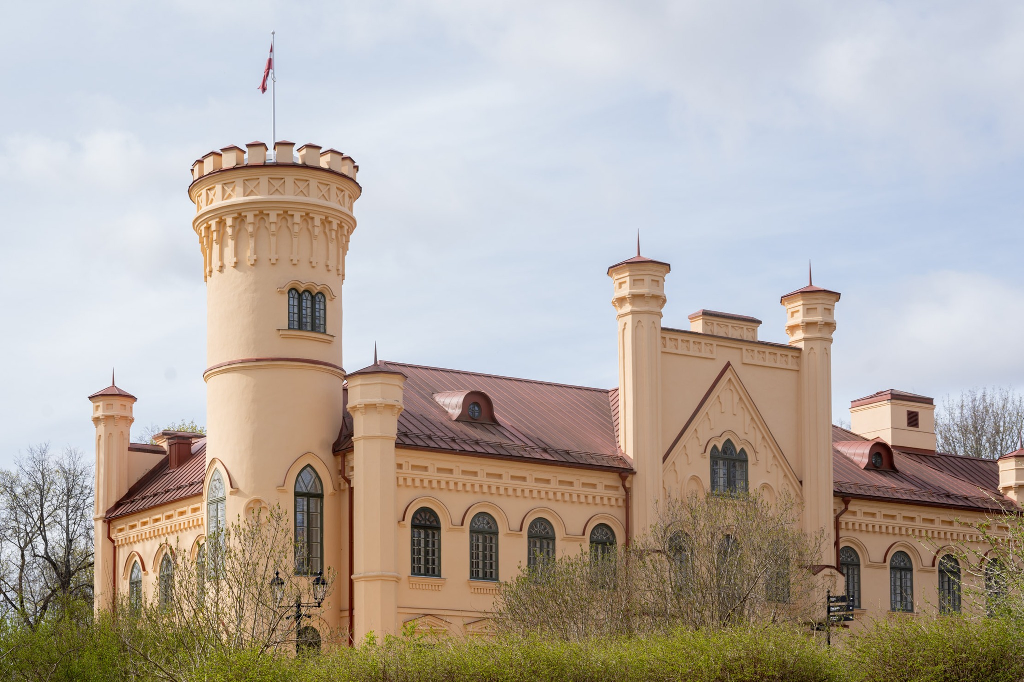 Historic castle with a turret and decorative architecture, surrounded by greenery against a cloudy sky. The building features arched windows and a prominent flag on top, showcasing its unique design and historical significance.