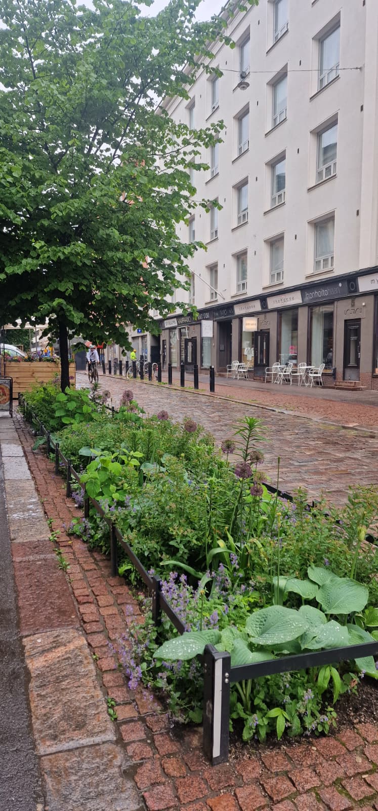 Lush greenery and flowering plants line a cobblestone street, with a large tree providing shade. Nearby, a row of outdoor café tables can be seen in front of modern buildings. The scene captures a vibrant urban landscape blending nature with city life.