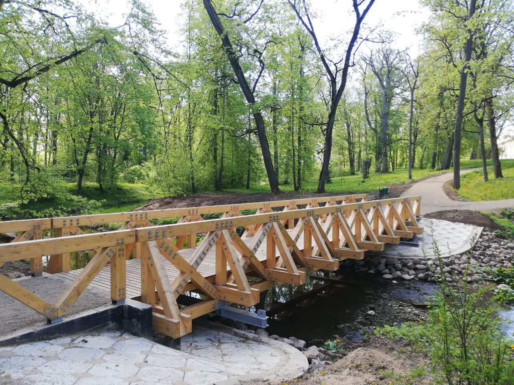 Wooden bridge crossing a small stream in a lush green forest, surrounded by trees and a walking path, showcasing nature and outdoor recreation.