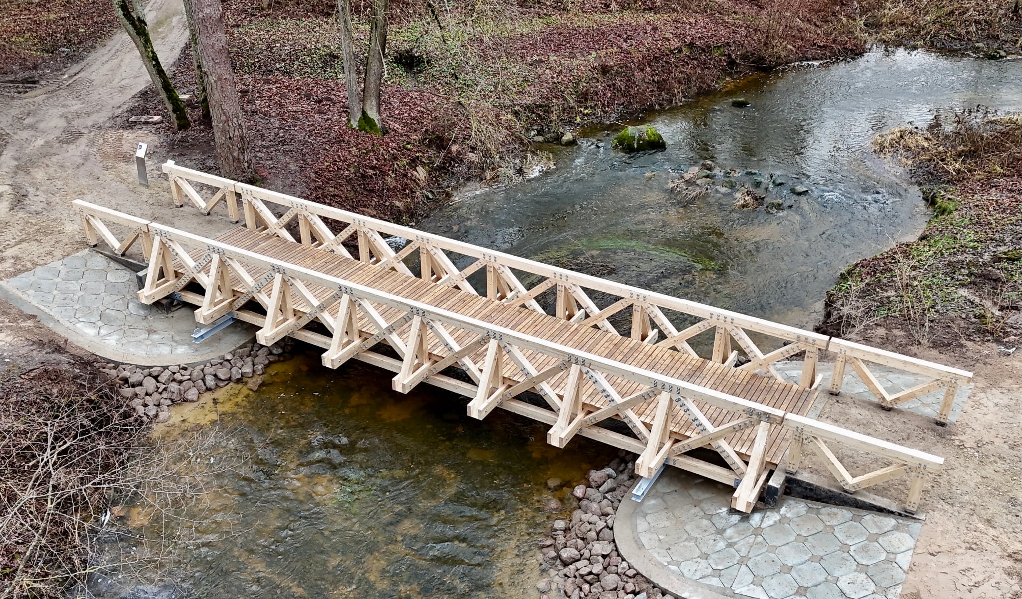 Wooden pedestrian bridge spanning a creek, surrounded by autumn foliage and a dirt path, showcasing modern design and natural integration.