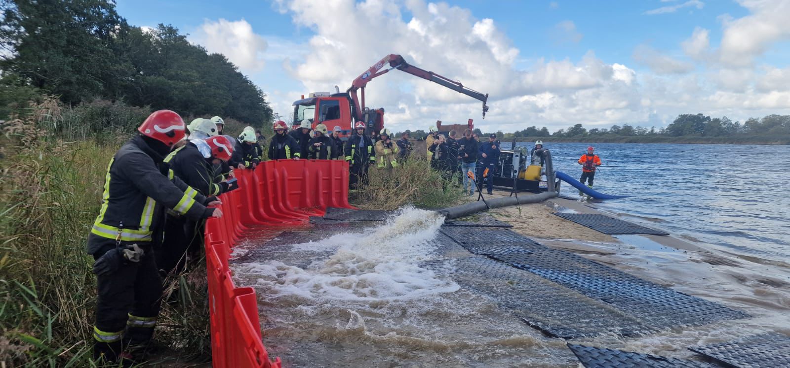 Firefighters and emergency personnel work together to manage a flood response operation, utilizing barriers and equipment near a riverbank, with a focus on safety and teamwork in a natural setting.