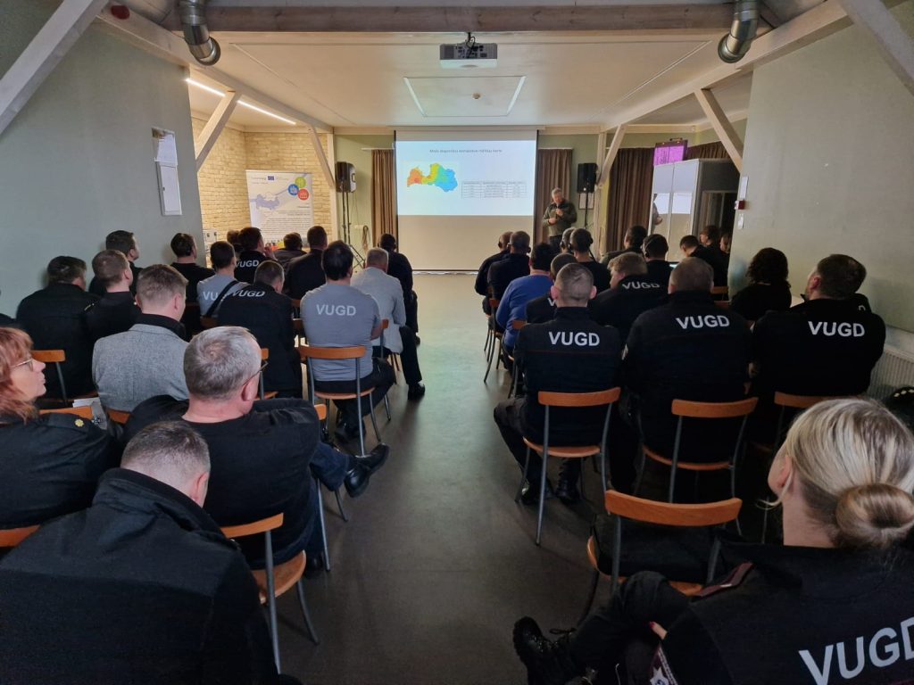 Audience attending a presentation at a conference, with attendees wearing VUGD uniforms, focused on a screen displaying a map and data analysis. The setting is a well-lit meeting room designed for professional discussions.