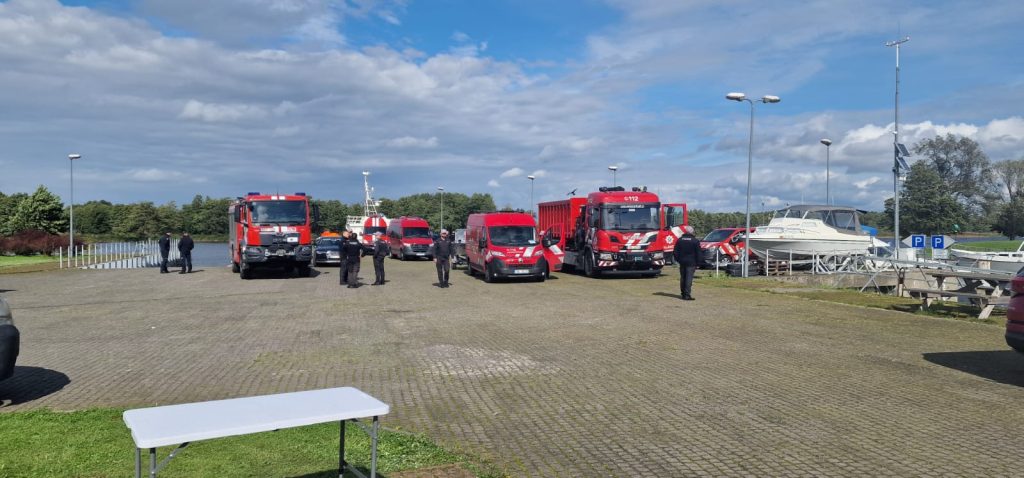 Emergency response vehicles and personnel gathered at a lakeside parking area, showcasing fire trucks and rescue units ready for action.