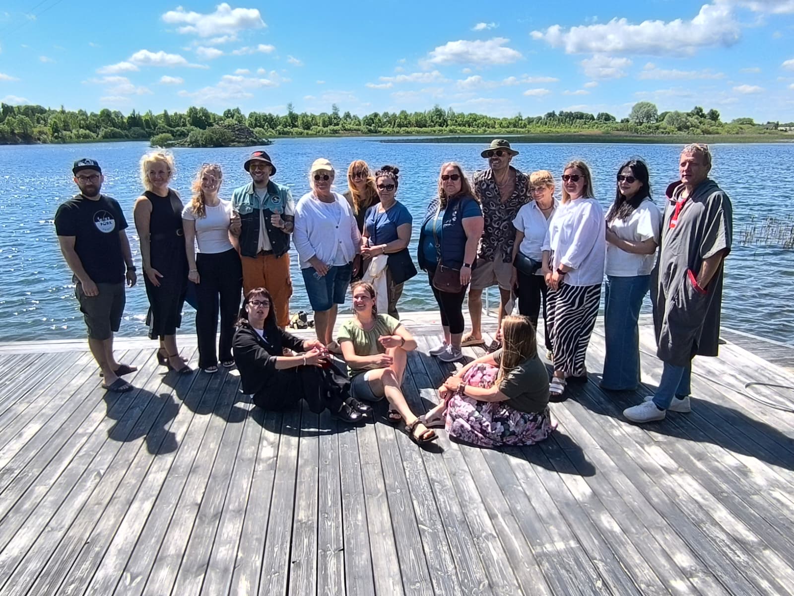 Group of diverse individuals posing together on a wooden dock by a lake, under a clear blue sky with fluffy clouds. The scene captures a summer outing, showcasing a mix of casual attire and happy expressions, surrounded by lush greenery in the background. Group of diverse individuals posing together on a wooden dock by a lake, under a clear blue sky with fluffy clouds. The scene captures a summer outing, showcasing a mix of casual attire and happy expressions, surrounded by lush greenery in the background.