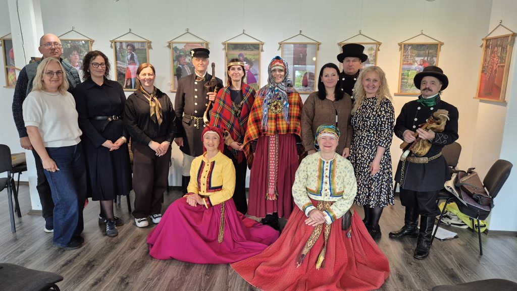 Group of people dressed in traditional costumes posing for a photo in an exhibition space, with framed artworks displayed on the walls. The scene captures a celebration of cultural heritage and community.