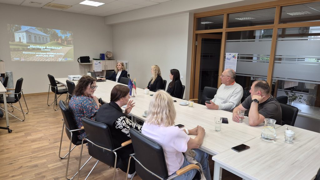 Group of professionals engaged in a discussion during the "Heritage Horizons" experience exchange meeting, featuring a presentation in the background. The meeting takes place in a modern conference room with a long table, chairs, and refreshments.