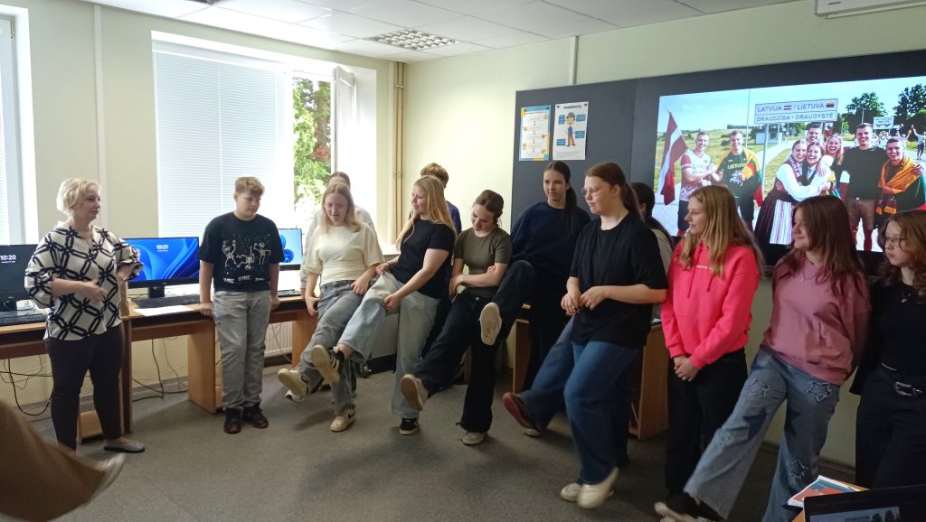 Group of students participating in an interactive classroom activity, with a teacher guiding them. The setting includes computers and educational materials, and a large screen displaying a cultural image from Latvia and Lithuania in the background.