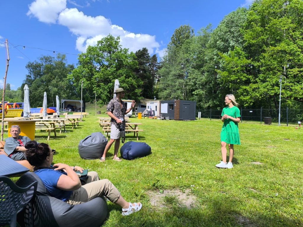 Outdoor gathering with people engaging in conversation on a sunny day, featuring picnic tables, bean bags, and lush greenery in the background. The scene includes a man wearing a patterned shirt and a hat speaking to a woman in a green dress, while others relax nearby. Outdoor gathering with people engaging in conversation on a sunny day, featuring picnic tables, bean bags, and lush greenery in the background. The scene includes a man wearing a patterned shirt and a hat speaking to a woman in a green dress, while others relax nearby.