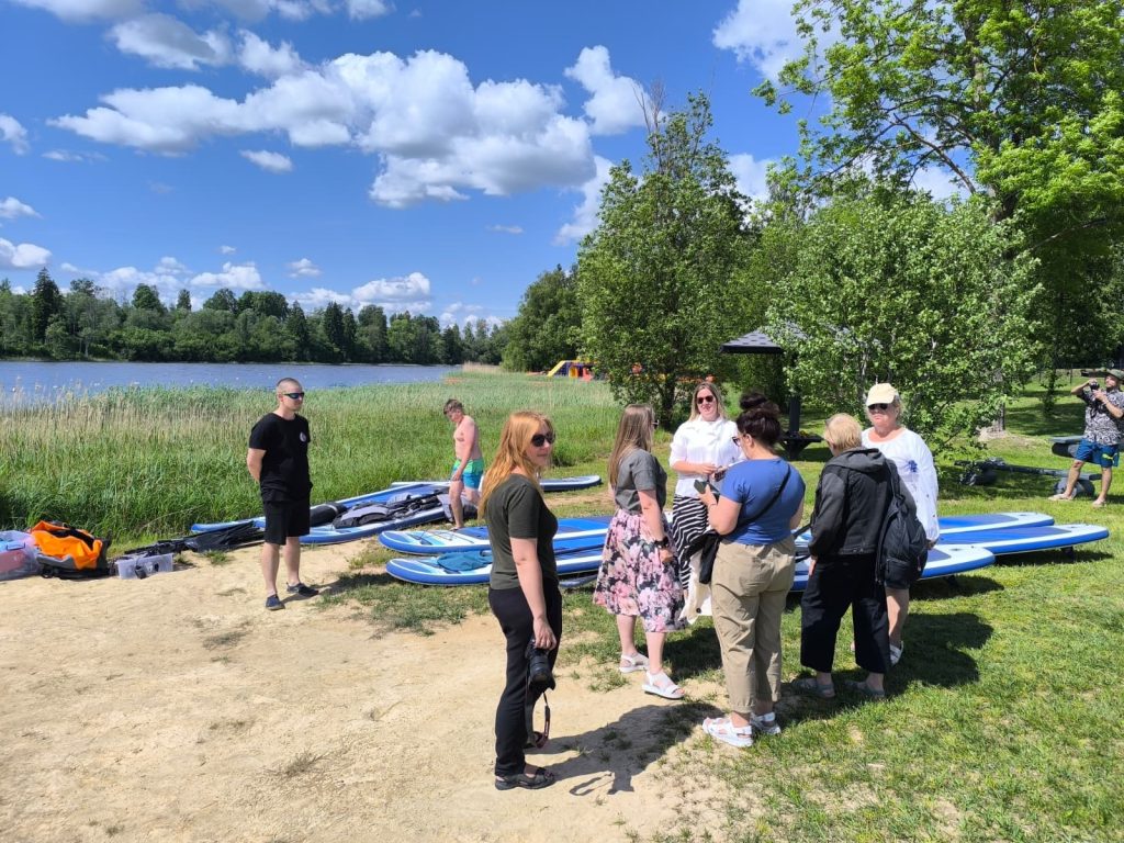 Group of people gathered by a serene lake, discussing paddleboarding options with several blue paddleboards visible in the foreground, surrounded by lush greenery and a clear blue sky. Group of people gathered by a serene lake, discussing paddleboarding options with several blue paddleboards visible in the foreground, surrounded by lush greenery and a clear blue sky.