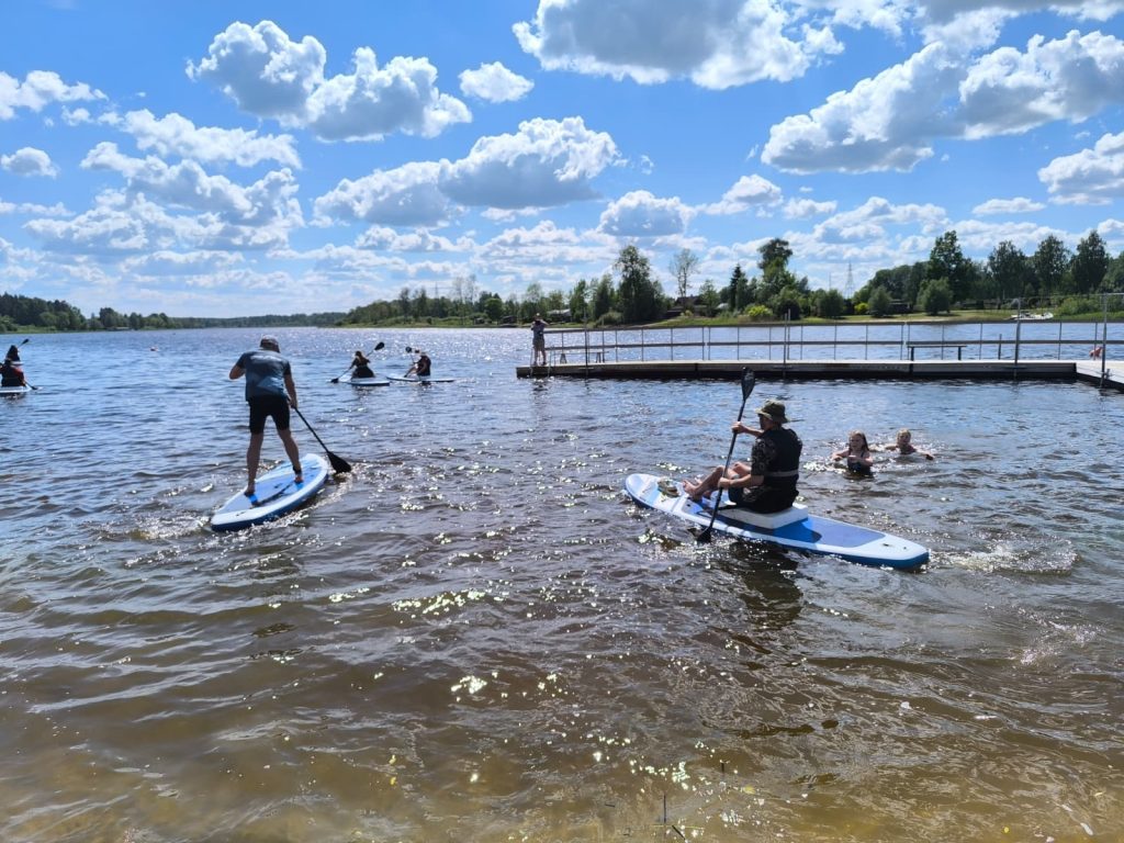 People enjoying paddleboarding on a sunny lake with a dock in the background, surrounded by trees and fluffy clouds in the sky. People enjoying paddleboarding on a sunny lake with a dock in the background, surrounded by trees and fluffy clouds in the sky.