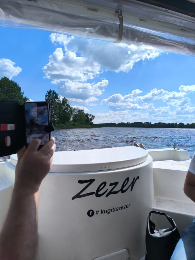 A person taking a photo with a smartphone while enjoying a scenic boat ride on a sunny day, with vibrant blue skies and fluffy clouds in the background. The boat features the name "Zezer" and is surrounded by lush greenery and calm waters. A person taking a photo with a smartphone while enjoying a scenic boat ride on a sunny day, with vibrant blue skies and fluffy clouds in the background. The boat features the name "Zezer" and is surrounded by lush greenery and calm waters.