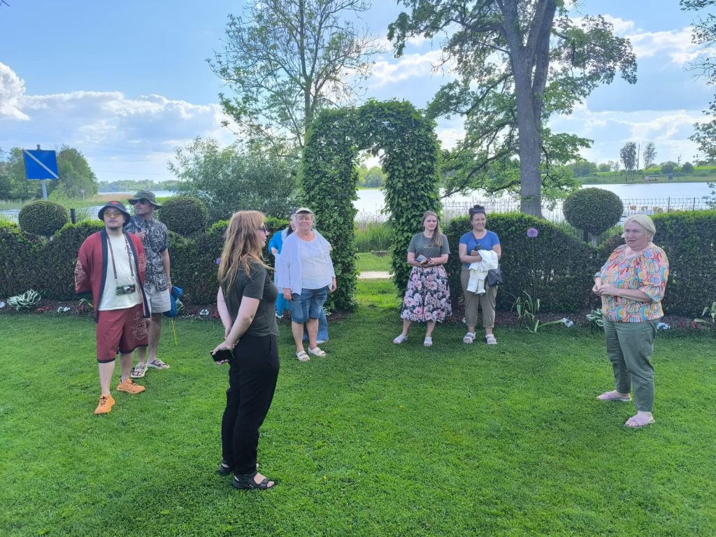 Group of people engaged in conversation outdoors by a lake, surrounded by lush greenery and ornamental hedges, under a partly cloudy sky. Group of people engaged in conversation outdoors by a lake, surrounded by lush greenery and ornamental hedges, under a partly cloudy sky.