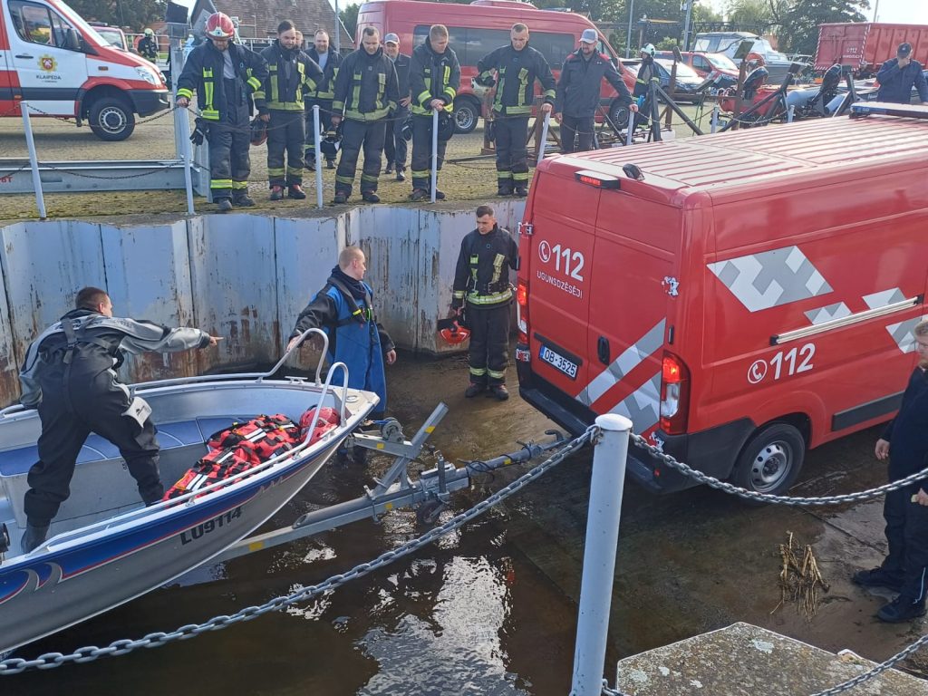 Emergency responders prepare a rescue boat for deployment at a waterway, with a red emergency vehicle in the background. Firefighters and rescue personnel in protective gear assist in the operation, ensuring safety measures are in place.