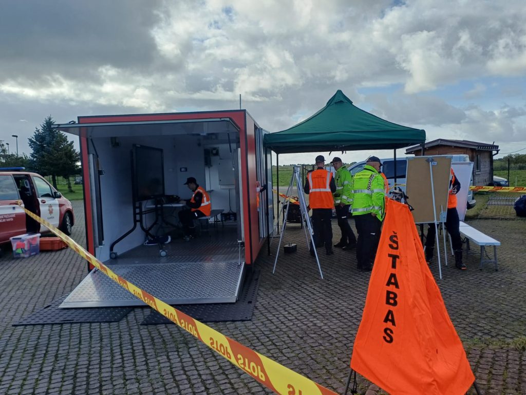 Emergency response team coordinating operations at a mobile command center, featuring a portable unit with a large screen, a green tent, and personnel in high-visibility jackets. The scene includes vehicles and equipment set up for effective emergency management.