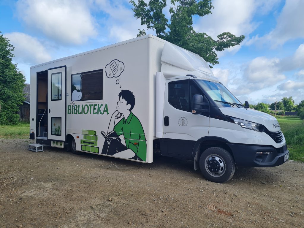 Mobile library truck with "Biblioteka" signage, featuring an illustration of a boy reading. The vehicle is parked in a grassy area with trees and a blue sky in the background.