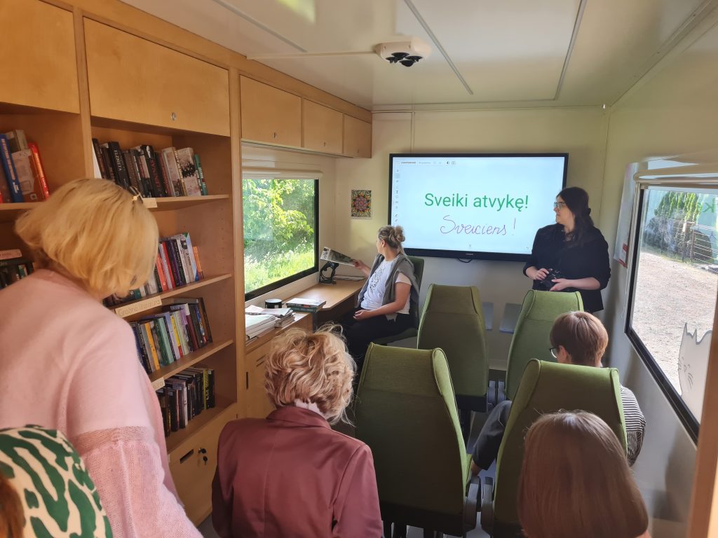 Interior of a mobile learning space featuring a presentation screen with welcoming text in Latvian. Attendees, including a speaker and seated participants, engage in a session surrounded by bookshelves filled with various titles. Natural light filters through the windows, enhancing the inviting atmosphere.
