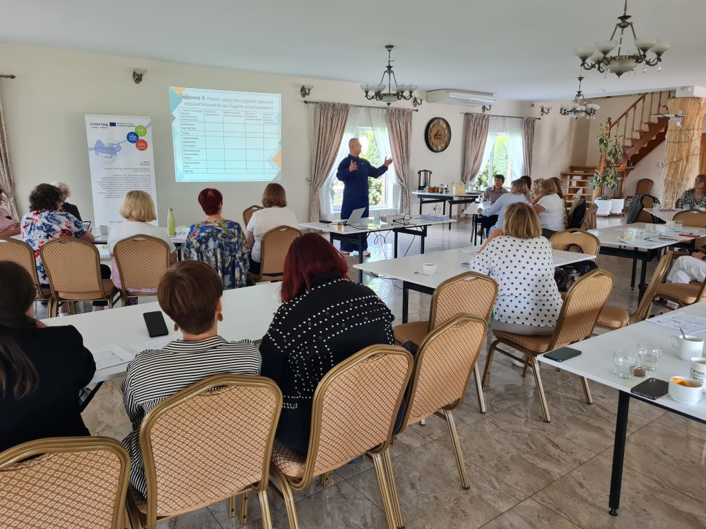 Audience participating in a workshop, with a speaker presenting in front of a screen displaying a table, in a well-lit conference room with tables set for discussion.