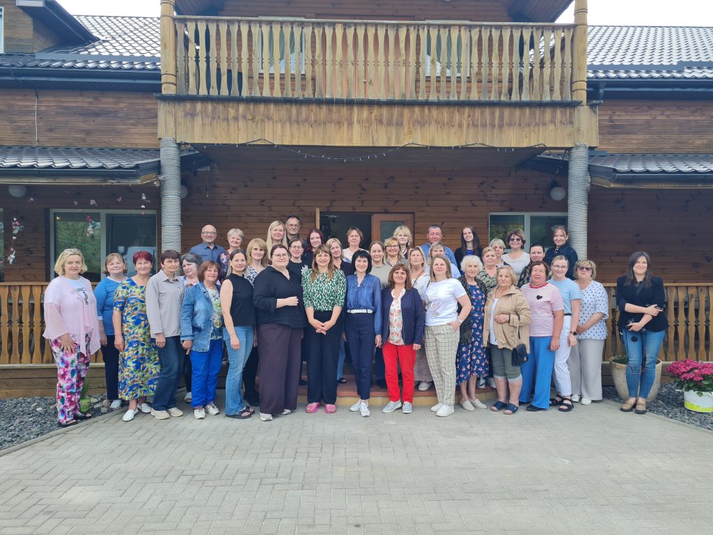 Group photo of a diverse gathering of approximately 40 individuals standing in front of a wooden building with a balcony, showcasing a friendly atmosphere and community spirit. The participants are dressed in casual attire, smiling and posing together on a paved area surrounded by plants.