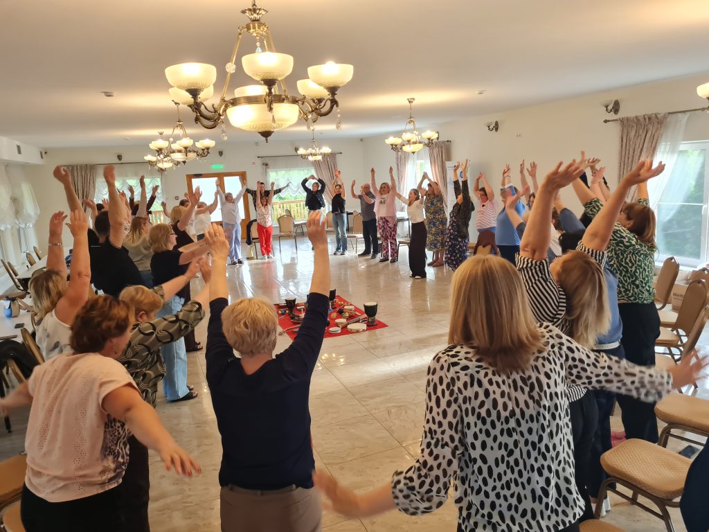 Group of women engaged in a wellness activity, raising their arms in a spacious, well-lit room with chandeliers. The setting features chairs arranged around a central area, suggesting a focus on movement and community interaction.