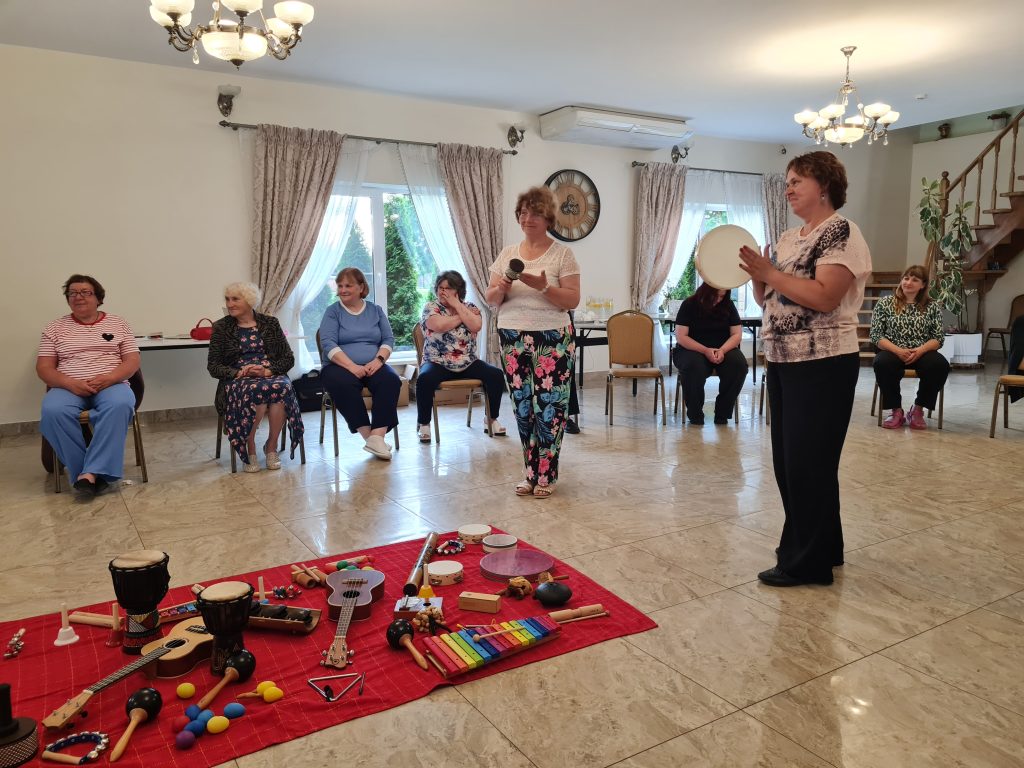 A group of women engaged in a music therapy session, sitting in a circle with various musical instruments displayed on a red mat. Participants are actively involved, with two women playing instruments while others observe and enjoy the session in a well-lit indoor space.