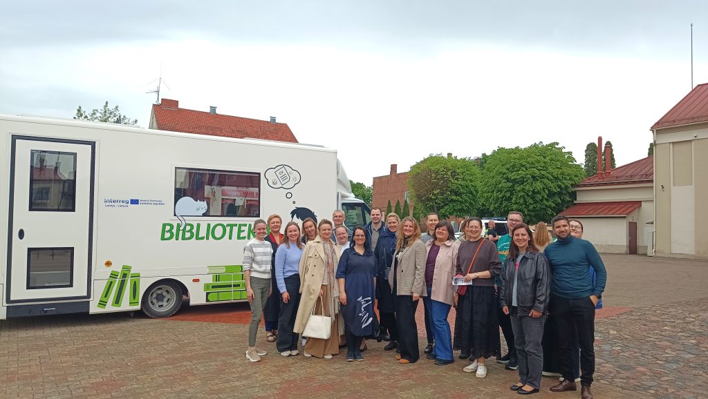 Group of people gathered in front of a mobile library (Bibliotēka) in Latvia, promoting literacy and community engagement. The vehicle features colorful graphics and is parked in a scenic outdoor setting.
