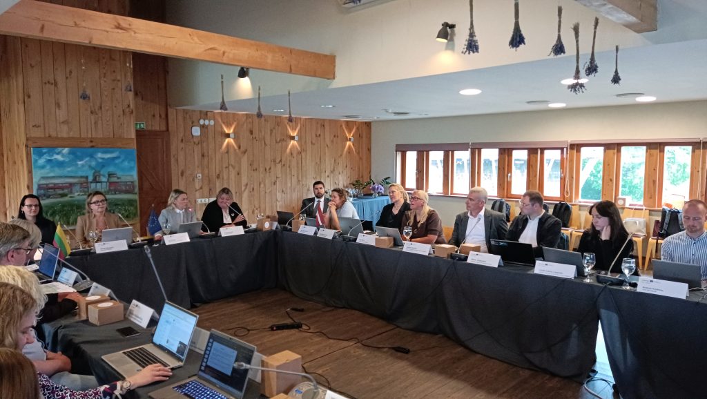 Group meeting in a conference room with wooden decor, featuring participants seated around a table with laptops and documents. Attendees engage in discussion, with flags and microphones visible, highlighting a collaborative environment. Natural light filters through windows, creating a warm atmosphere.