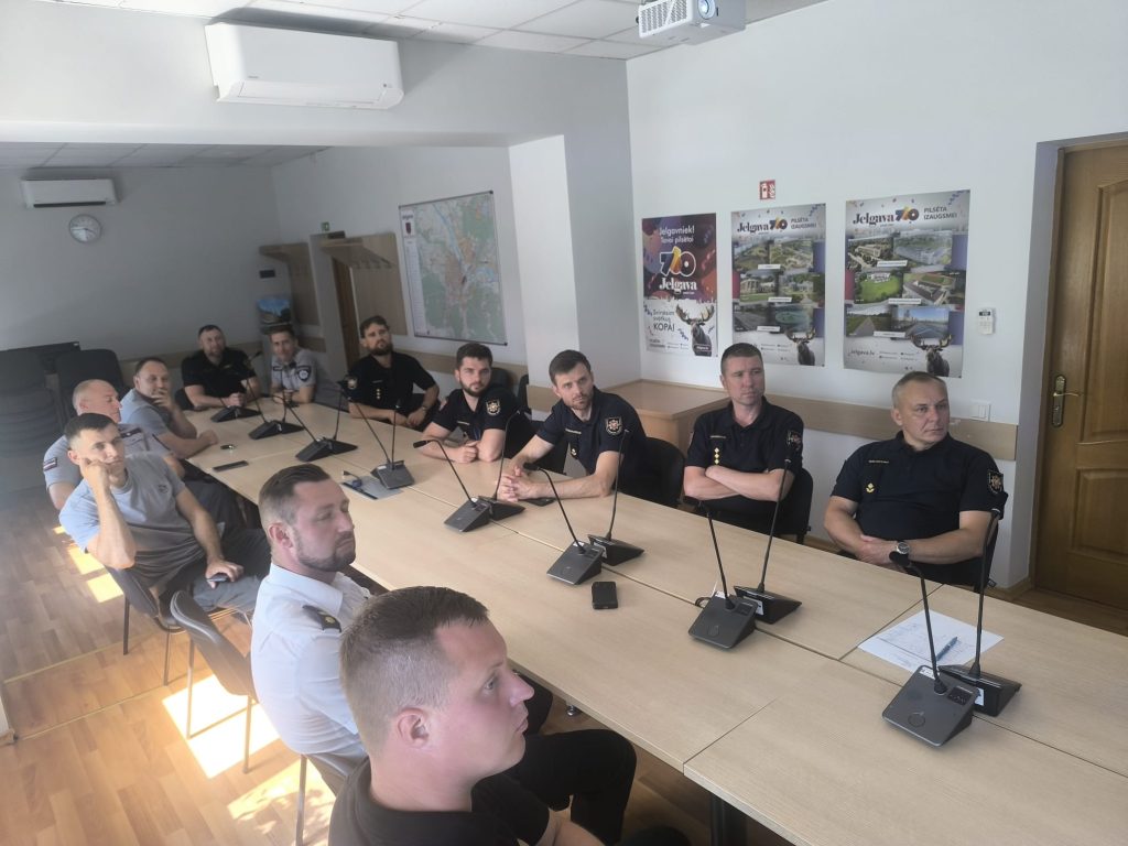 Group of law enforcement officers attending a meeting in a conference room, featuring a long table with microphones and a map on the wall. Several posters related to local attractions are visible in the background, indicating a focus on community engagement and public safety.