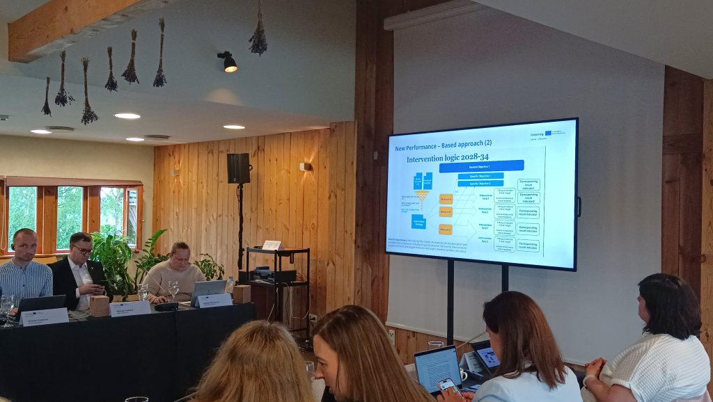 Meeting participants engage in a discussion while reviewing a presentation on the "New Performance-Based Approach" and "Intervention Logic 2028-34," displayed on a screen in a wooden-paneled conference room.