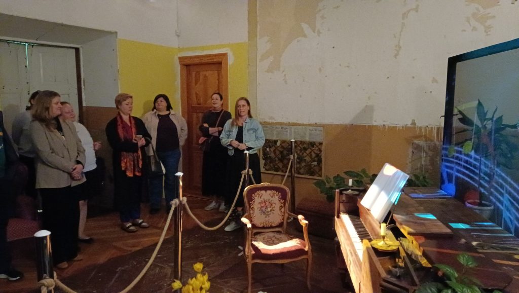 Group of visitors observing an art installation in a rustic room, featuring a vintage piano and a colorful projection on a screen, with yellow and peeling walls, and decorative plants nearby.