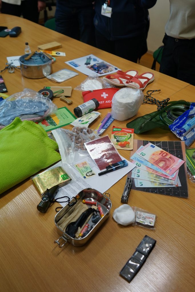Assorted items displayed on a wooden table, including a first aid kit, cash in various denominations, cooking equipment, a green blanket, personal identification, and various tools and supplies, highlighting emergency preparedness and survival gear.