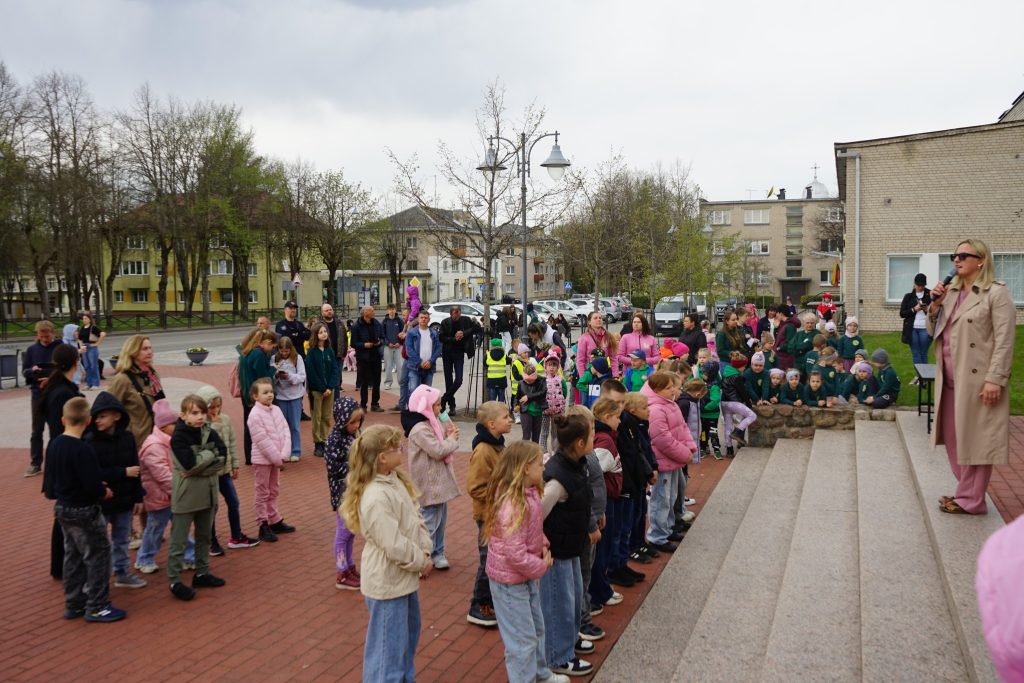 A crowd of children and adults gathers in a public square, with a woman speaking on steps in the foreground. The scene captures a community event, showcasing children in colorful clothing and engaged participants, set against a backdrop of residential buildings and trees.