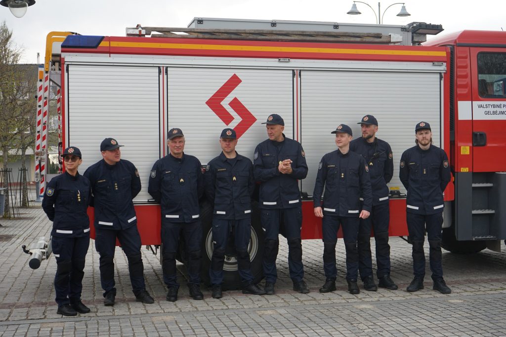 Group of firefighters in uniform standing in front of a fire truck, showcasing teamwork and readiness for emergency response. The background features a modern fire station setting.