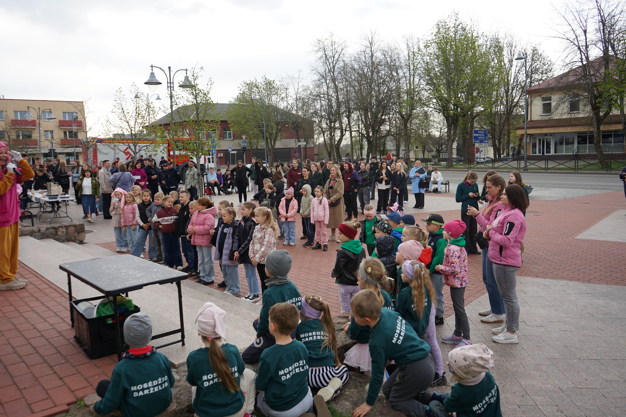 A lively outdoor event featuring a clown entertaining a large crowd, including many children dressed in colorful clothing, while parents and adults watch from the background. The setting includes trees and buildings, creating a community atmosphere.