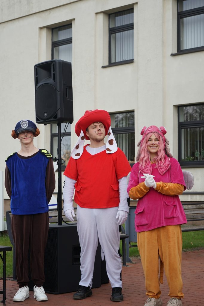 Three individuals dressed in colorful cartoon character costumes stand outdoors in front of a speaker. The first person on the left wears a blue and brown outfit with a police hat, the middle person is in a red and white dog costume, and the third person on the right is dressed in a pink outfit with animal ears. The background features a building with large windows.