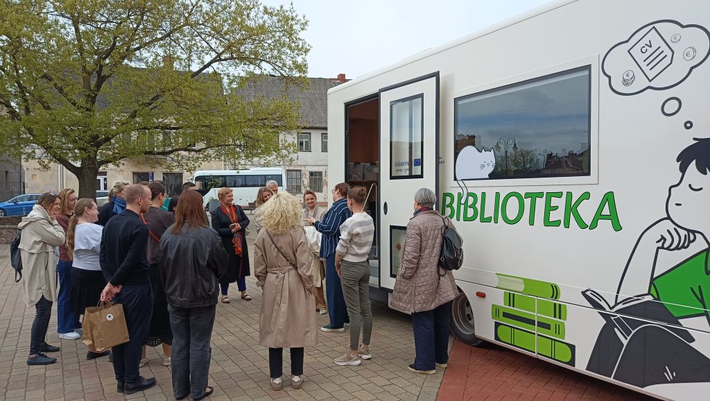 A diverse group of people gathers around a mobile library truck, labeled "BIBLIOTEKA," in an outdoor setting, engaging in conversation and activities related to reading and community resources.