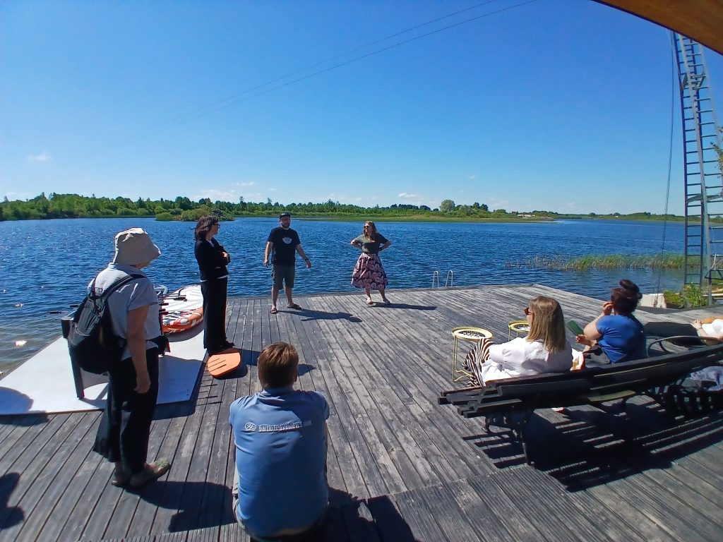 Group of people engaged in a workshop on a wooden deck by a serene lake under a clear blue sky, with some participants seated and others actively participating in discussions and activities. Group of people engaged in a workshop on a wooden deck by a serene lake under a clear blue sky, with some participants seated and others actively participating in discussions and activities.
