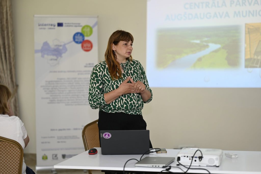 A woman with long brown hair, wearing a green patterned blouse, presents information about the Central Administration of Augšdaugava Municipality at a conference. A laptop and projector are set up on a table, with a background display showing images of landscapes and a map.