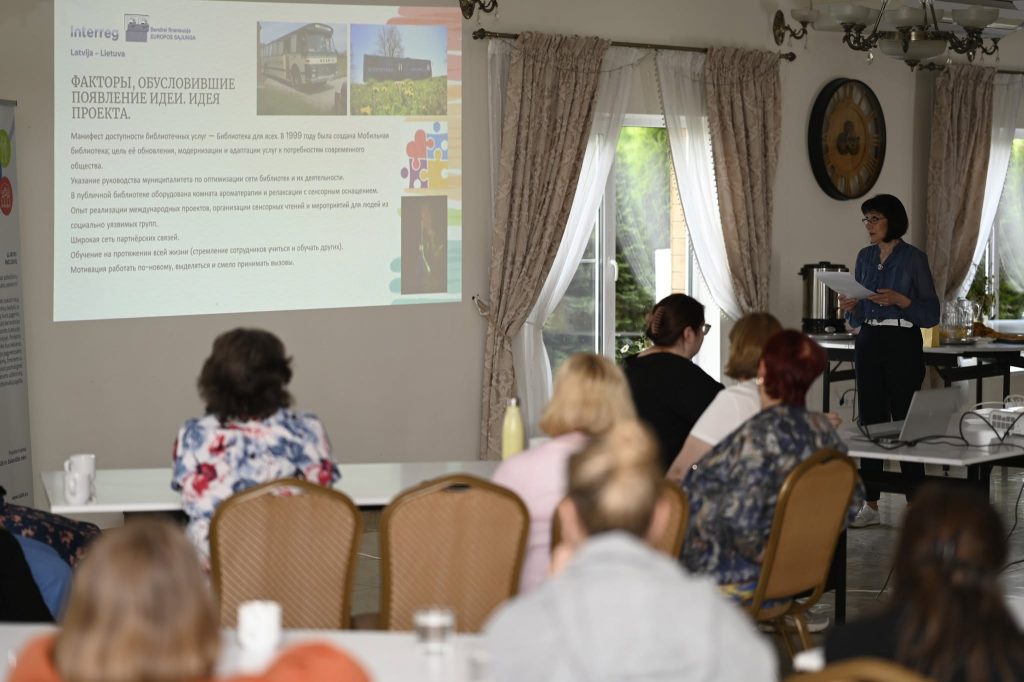 Presentation on project ideas and factors influencing development, with a speaker addressing an audience in a conference setting. A projector displays information in the background, while attendees engage and take notes. The environment features natural light and a professional setup, emphasizing collaboration and learning.