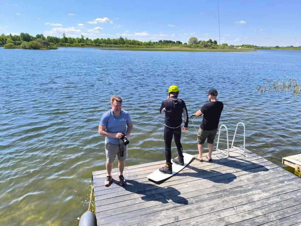 Three men prepare for water sports at a lakeside dock on a sunny day. One man stands with a camera, while another is suited for wakeboarding, and a third is holding a cable. The calm water and lush greenery in the background create a vibrant outdoor setting. Three men prepare for water sports at a lakeside dock on a sunny day. One man stands with a camera, while another is suited for wakeboarding, and a third is holding a cable. The calm water and lush greenery in the background create a vibrant outdoor setting.