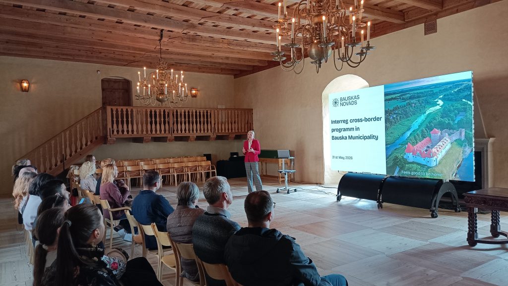 Presentation on the Interreg cross-border program in Bauska Municipality, featuring a speaker in a red jacket addressing an audience in a wooden hall with a large screen displaying program details and an aerial view of Bauska Castle.